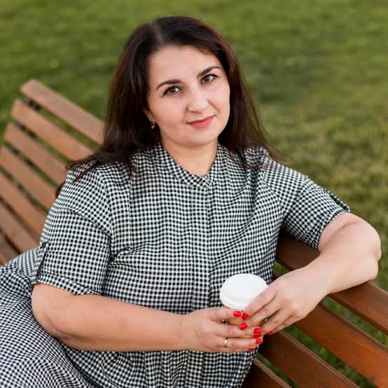 Woman seated on a park bench holding a white takeaway coffee cup, wearing a checkered dress.