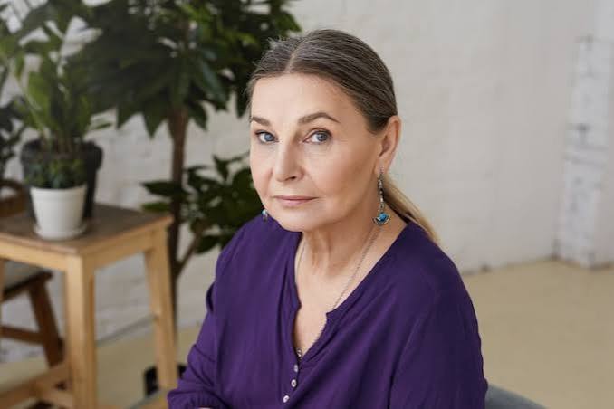 Portrait of a mature woman in a purple blouse, seated indoors with plants in the background.