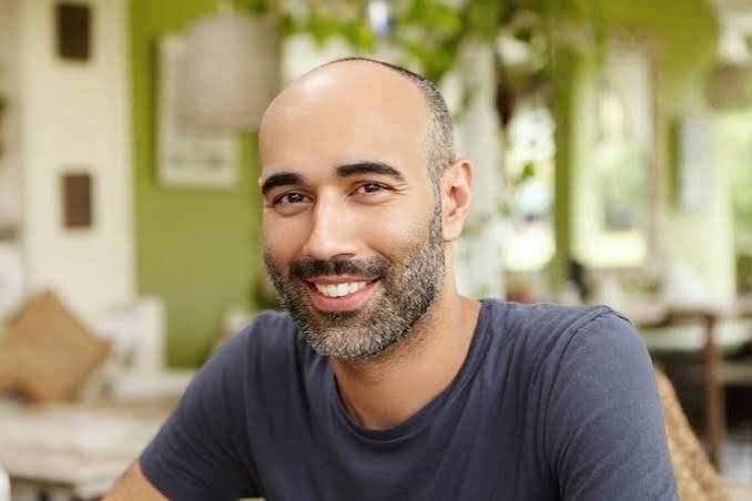 Smiling man with a shaved head and beard sits at a cozy cafe, wearing a dark blue t‑shirt and looking at the camera.